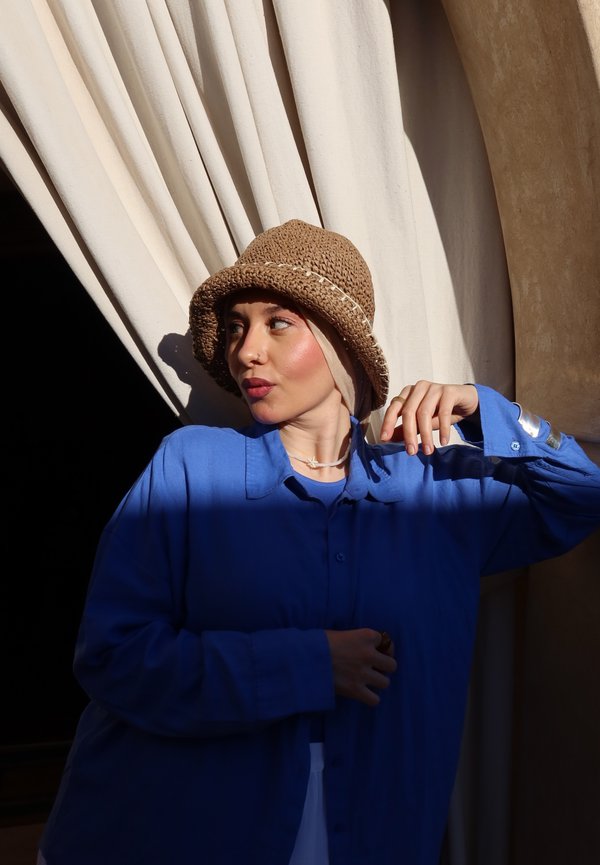 Femme avec chapeau marron clair et chemise bleue pose à côté d'un rideau en tissu clair sous une lumière chaude du soleil, regardant vers la droite avec la main levée près du visage.
