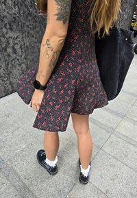 Μαύρο φόρεμα με λουλούδια σε κόκκινο χρώμα και λευκές βούλες; φαρδιά φούστα. Μαύρα παπούτσια με λευκές κάλτσες. Ορατό τατουάζ στο χέρι. Σκούρα τσάντα.