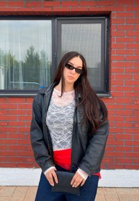 Čierna kožená bunda cez biely čipkovaný top a červené tričko, doplnené tmavými džínsami a čiernym klobúkom. Nosené slnečné okuliare.