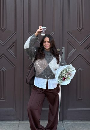 Joven mujer con suéter estampado y pantalones marrones sosteniendo un ramo en una mano y levantando una taza de café con la otra, de pie frente a grandes puertas marrones.