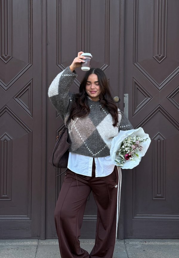 Młoda kobieta w wzorzysty sweter i brązowych spodniach trzyma bukiet w jednej ręce, a drugą podnosi filiżankę kawy, stoi przed dużymi brązowymi drzwiami.