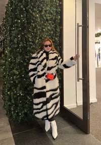 Femme portant un manteau en fausse fourrure aux rayures zébrées noires et blanches, des bottes blanches et des lunettes de soleil, debout près d'une porte vitrée avec des guirlandes lumineuses sur une verdure derrière elle.