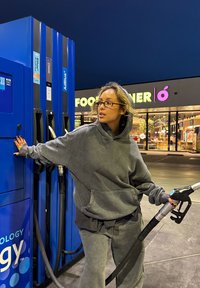 Felpa grigia con cappuccio e pantaloni abbinati, che interagisce con una pompa di benzina blu. L'area circostante include un negozio ben illuminato sullo sfondo.