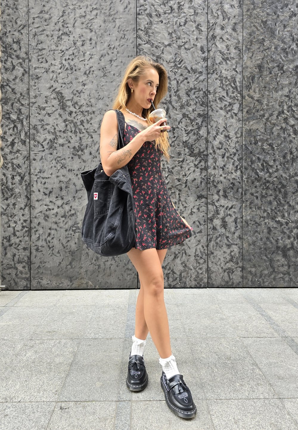 Μαύρο φλοράλ φόρεμα με ρυθμιζόμενους ιμάντες, συνδυασμένο με μια μαύρη τσάντα tote. Μαύρα παπούτσια με λευκές κάλτσες. Λείο, γκρίζο υφασμάτινο φόντο.