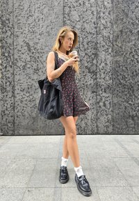 Μαύρο φλοράλ φόρεμα με ρυθμιζόμενους ιμάντες, συνδυασμένο με μια μαύρη τσάντα tote. Μαύρα παπούτσια με λευκές κάλτσες. Λείο, γκρίζο υφασμάτινο φόντο.