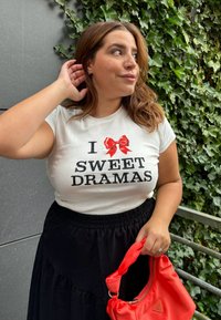 Hvid T-shirt med rød sløjfegrafik og sort tekst "I SWEET DRAMAS". Sort nederdel med elastisk talje. Holder en rød stof skuldertaske.
