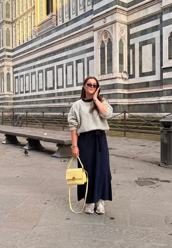 Femme avec des lunettes de soleil, un pull gris, une jupe bleu marine et des baskets tenant un sac à main jaune, debout près d'un bâtiment historique orné avec une façade en pierre rayée.