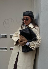 Trench-coat couleur crème, pull gris, pochette noire tissée, gants marron et casquette en cuir noir ornée de clous argentés, posant contre un mur.