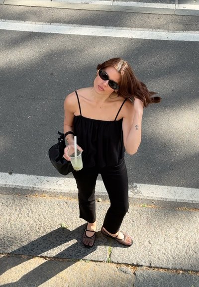 Mujer con ropa negra y gafas de sol está en el borde de la calle, sosteniendo una bebida con pajita y ajustándose el cabello con una mano.