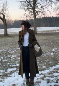 Γυναίκα με σκούρο πράσινο παλτό, μαύρο καπέλο και γάντια, στέκεται σε χιονισμένο έδαφος με άγρια δέντρα και δάσος στο φόντο κατά το σούρουπο.