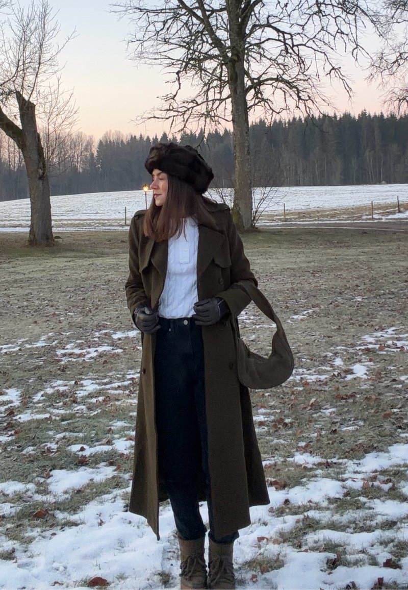 Ženska v temno zeleni jakni, črnem klobuku in rokavicah stoji na zasneženi zemlji s poganjajočimi drevesi in gozdom v ozadju ob mraku.