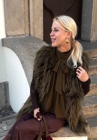 Lažni krzneni prsluk u maslinastoj zelenoj boji preko tamnosmeđe bluze s mašnom, uparen s smešnim hlačama. Zlate kružne naušnice i prsten kao akcent.