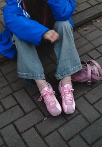 Ροζ αθλητικά παπούτσια με κορδόνια, λεία υφή. Φοριούνται με ανοιχτό μπεζ κάλτσες και ανοιχτό μπλε φαρδιά παντελόνια. Μωβ τσ άντα κοντά σε πλακόστρωτο έδαφος.