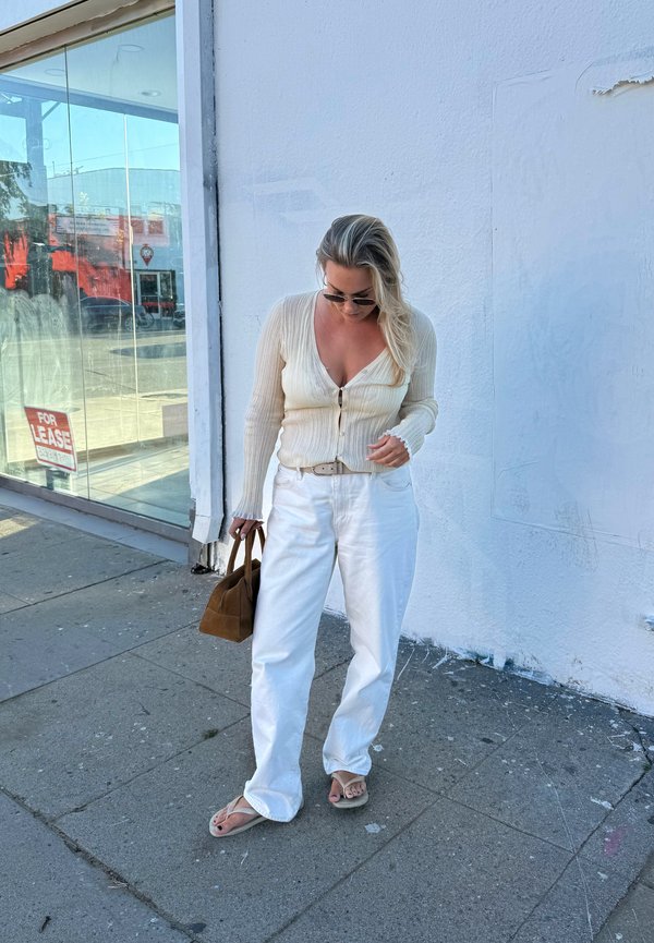 Frau mit Sonnenbrille trägt eine cremefarbene Strickjacke, weiße Hose und Sandalen, hält eine braune Tasche und steht auf einem Bürgersteig neben einer weißen Wand.