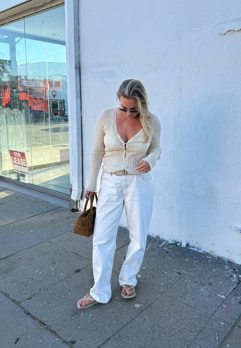 Donna con occhiali da sole che indossa un cardigan crema, pantaloni bianchi e sandali, con una borsa marrone, in piedi su un marciapiede accanto a un muro bianco.