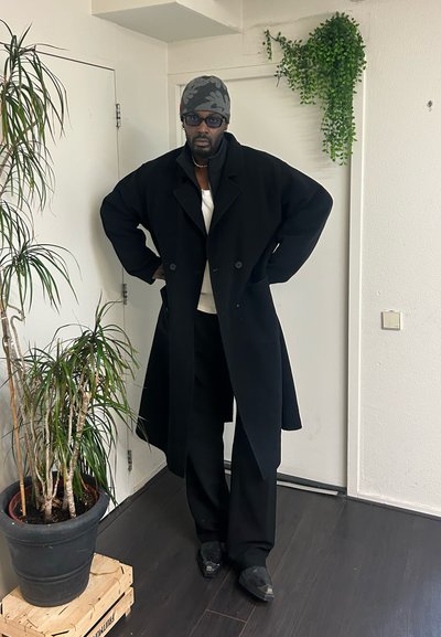 Abrigo negro de doble botonadura con silueta oversize, usado sobre una blusa blanca; combinado con pantalones negros y zapatos negros. Entorno interior con plantas.