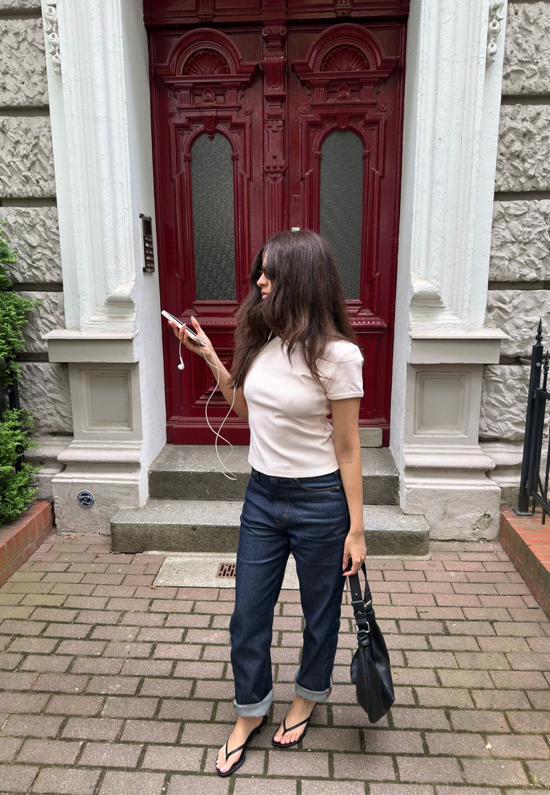 Mujer con camisa blanca y vaqueros remangados de pie sobre pavimento de ladrillo, sosteniendo un teléfono con auriculares y un bolso negro cerca de una puerta roja.