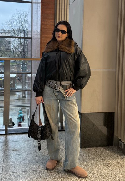 Mujer con chaqueta negra con cuello de piel sintética, vaqueros azul claro, zapatos marrones, gafas de sol negras, llevando bolso negro en el interior junto a la ventana.