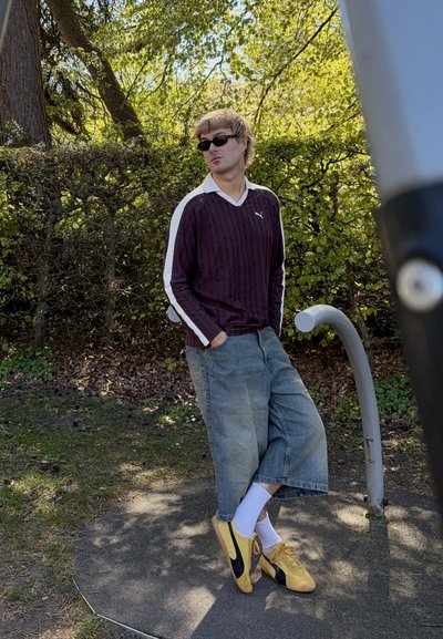 Joven con gafas de sol, suéter púrpura de Puma, pantalones cortos anchos de mezclilla y zapatillas amarillas, de pie con las manos en los bolsillos cerca de un seto al aire libre.