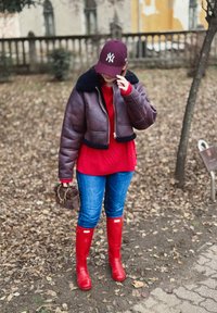 Jachetă maro scurtă cu guler din blană neagră, pulover roșu cu model împletit, blugi albaștri și cizme de cauciuc roșii. Geantă maro cu lanț auriu.