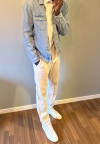 Veste en denim bleu clair sur une chemise blanche, associée à un pantalon blanc ample et des baskets blanches. Fond en parquet en bois.