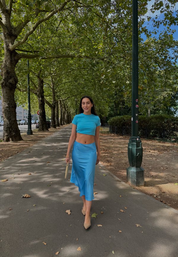 Femme en haut court bleu et jupe marchant sur un trottoir bordé d’arbres, tenant une pochette beige par une journée ensoleillée.