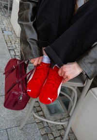 Raudoni Puma sportbačiai su baltais akcentais, pagaminti iš zomšos, su raišteliais. Ant kėdės, šalia žmogaus su juodomis kelnėmis, guli burgundiška rankinė.