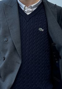 Κοντομάνικος ναυτικός πουλόβερ με πλέξη καλωδίων κάτω από ένα γκρι σακάκι διπλής όψεως. Εντυπωσιακό πράσινο λογότυπο κροκοδείλου πάνω στο πουλόβερ. Ορατό λευκό πουκάμισο με γιακά.