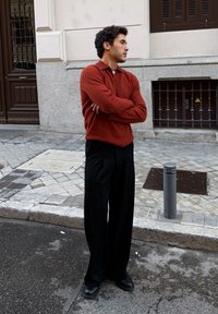Pull tricot texturé rouge avec col, associé à un pantalon large noir et des chaussures noires, debout sur une rue pavée.
