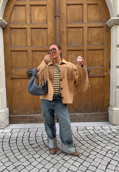 Mujer con chaqueta marrón con flecos y gafas de sol comiendo gelato, parada en una calle empedrada frente a grandes puertas dobles de madera.