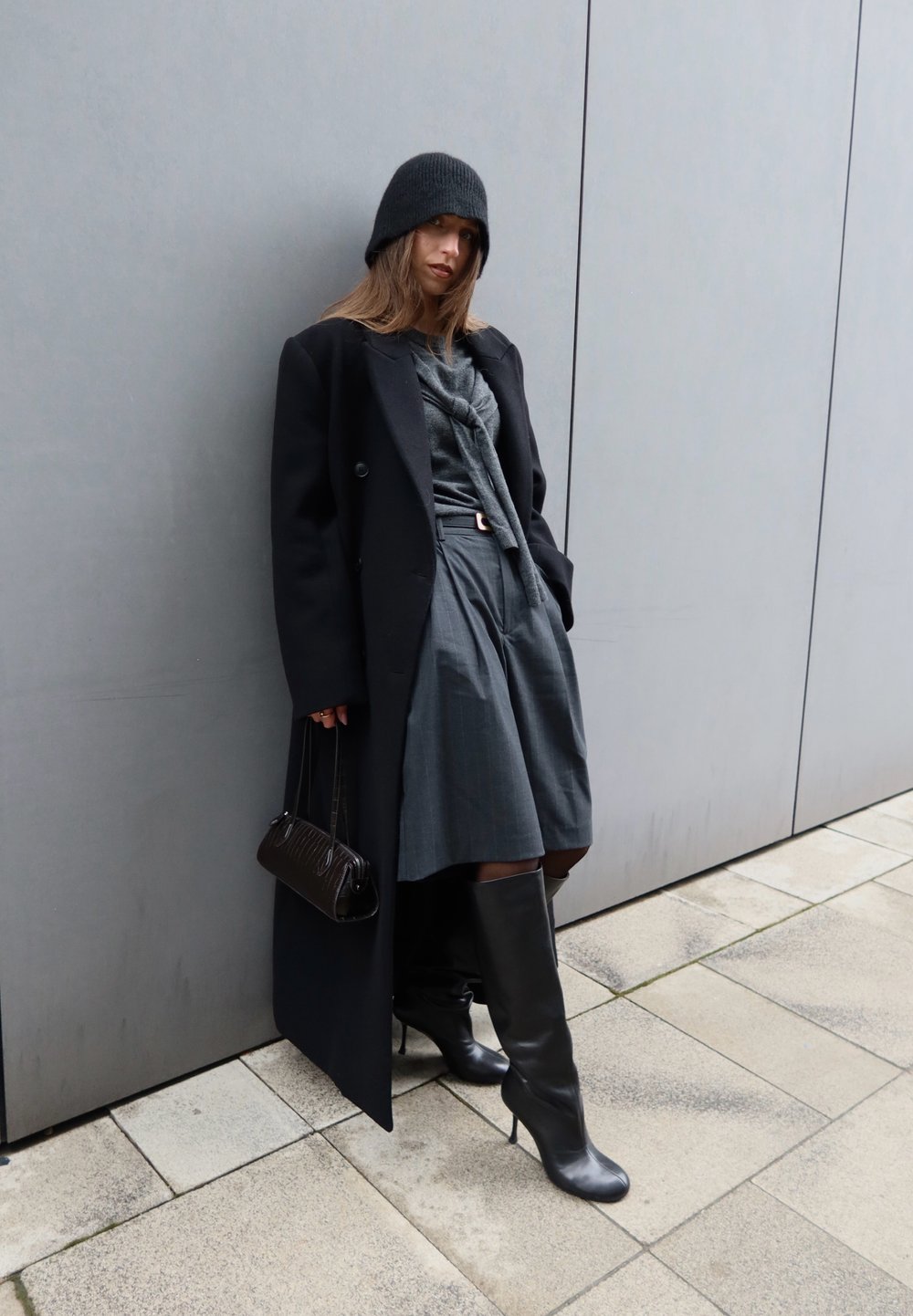 Femme portant un bonnet noir, un long manteau noir, un haut gris et une jupe, des bottes noires montant jusqu'aux genoux, tenant un petit sac à main, appuyée contre un mur gris en extérieur.
