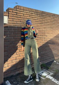 Άτομο που φοράει πουλόβερ με ουράνιο τόξο ρίγες, χακί παντελόνι, κασκόλ και μπλε καπέλο, στέκεται δίπλα σε ηλιόλουστο τοίχο από τούβλα σε εξωτερικό χώρο στάθμευσης.