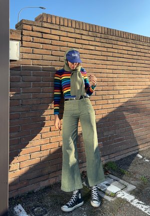 Persona con un suéter a rayas de colores del arcoíris, pantalones verde oliva, bufanda y gorra azul, de pie contra una pared de ladrillos iluminada por el sol en un área de estacionamiento al aire libre.