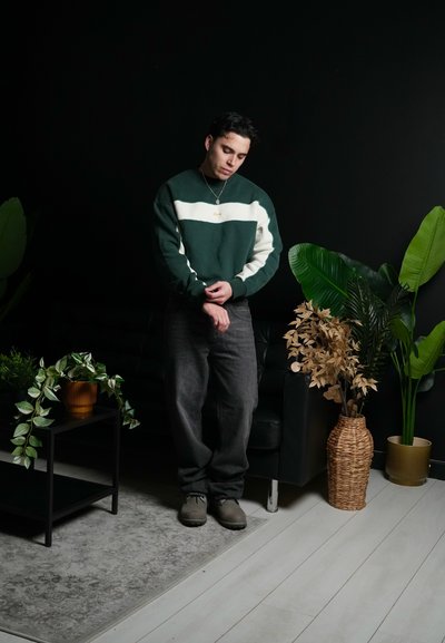 Joven ajustándose la manga, de pie junto a un sofá negro y una mesa con plantas en macetas en una habitación con pared oscura y suelo de madera clara.