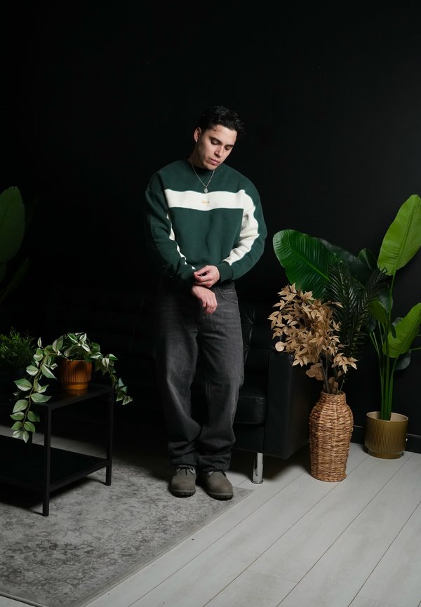 Noor mees sättimas varrukat, seistes musta diivani ja potitaimedega laua kõrval toas, kus on tume sein ja hele puidust põrand.