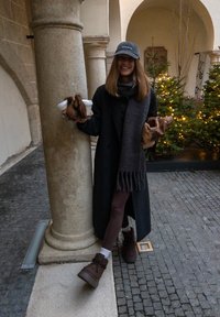 Femme portant un manteau sombre et une écharpe, tenant des tasses de café et des pâtisseries, appuyée contre une colonne en pierre dans une cour décorée avec des sapins de Noël illuminés.