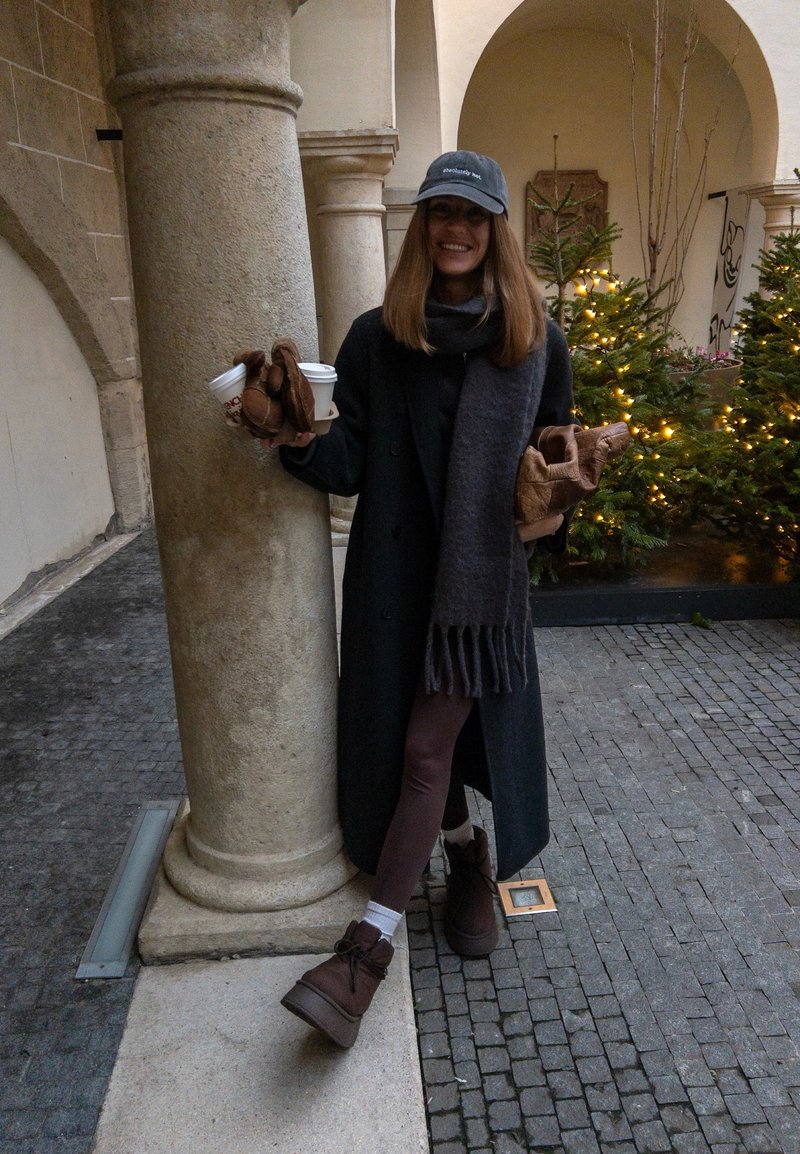 Frau in dunklem Mantel und Schal, hält Kaffeetassen und Gebäck, lehnt an einer Steinsäule in einem dekorierten Hof mit beleuchteten Weihnachtsbäumen.