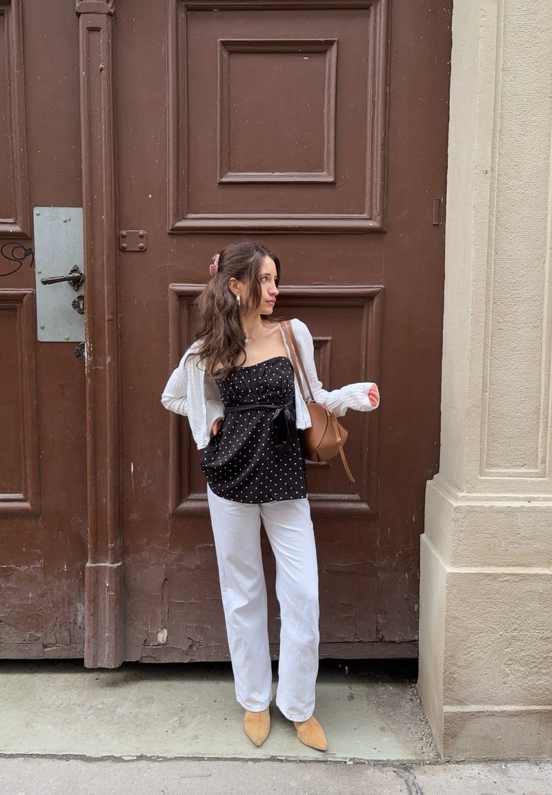 Pitkätukkainen nainen seisoo suuren ruskean puuoven edessä, pukeutuneena mustaan pilkulliseen paitaan, valkoisiin housuihin, beigen värisiin kenkiin ja ruskeaan olkalaukkuun.