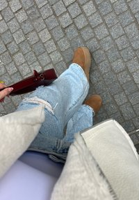 Ανοιχτό μπλε σκισμένα τζιν, καφέ μπότες με τακούνι και μια κόκκινη τσάντα. Η επιφάνεια είναι από cobblestone, με λεπτομέρειες σε κοντινό πλάνο για τα υφάσματα και τις υφές.