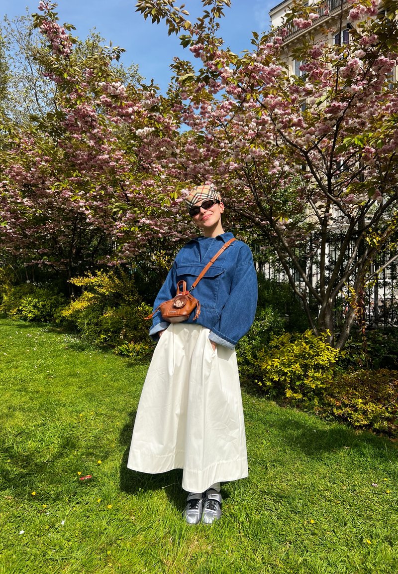 Persona che indossa una giacca di jeans, gonna bianca, berretto fantasia, occhiali da sole e borsa a tracolla marrone in piedi sull'erba sotto ciliegi rosa in fiore.