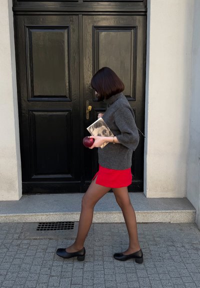 Mujer con un suéter gris y falda roja camina frente a unas puertas dobles negras, sosteniendo un libro y una manzana roja en sus manos.