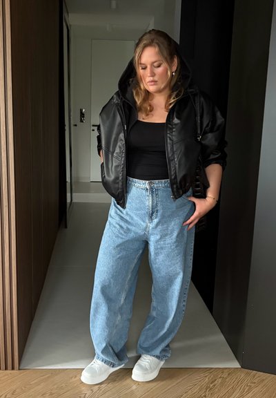 Chaqueta acolchada negra con capucha, camiseta negra sin mangas, jeans de pierna ancha en azul claro y zapatillas blancas. Materiales suaves y corte relajado.