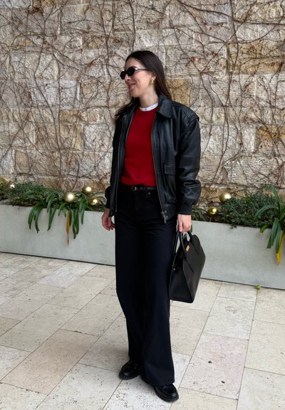 Mujer con chaqueta de cuero negra, suéter rojo, pantalones negros y zapatos, sosteniendo un bolso negro, de pie en el interior junto a una pared de piedra y plantas.