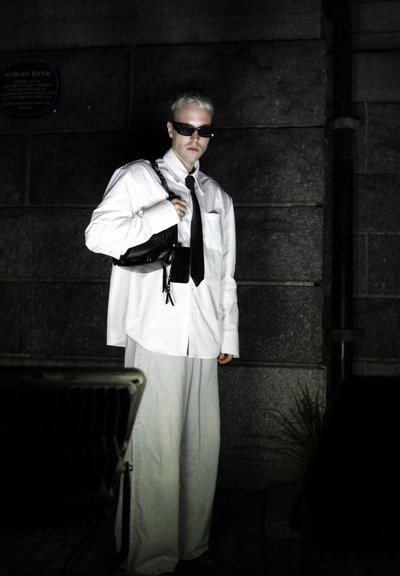 Camisa blanca de gran tamaño, corbata negra, bolso y gafas de sol; pantalones grises de pierna ancha; fondo oscuro texturizado; iluminación dramática que acentúa los rasgos.