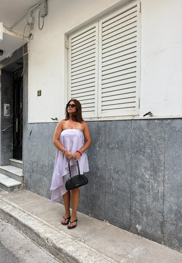 Femme en robe lilas sans bretelles et lunettes de soleil noires tenant un sac à main noir debout sur un trottoir à côté d’un mur de bâtiment blanc et gris.