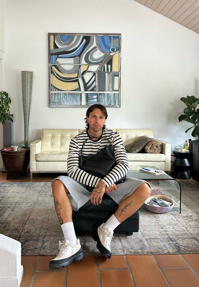 Bolso de cuero negro, camiseta de rayas de manga larga, pantalones cortos grises, calcetines blancos y zapatillas deportivas. Interior moderno con arte mural abstracto y plantas.