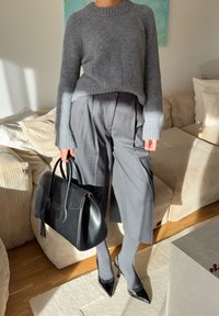 Grauer, kurzer Pullover mit geripptem Saum, hoch taillierte graue Plisseehose, schwarze Handtasche, graue Socken und schwarze Lack-High Heels.