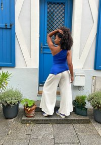 Top blu con spalla scoperta, pantaloni bianchi a gamba larga e sneaker blu; in piedi su gradini di pietra con piante in vaso e uno sfondo di porta blu.