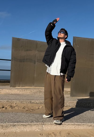 Chaqueta acolchada negra, camisa blanca a cuadros, pantalones anchos marrones, guantes sin dedos y gafas de sol, de pie sobre una superficie arenosa.