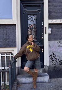 Felpa marrone con testo "Wyoming", pantaloni short in denim grigi e stivali color sabbia al ginocchio. Porta nera con lavorazione in ferro decorativa; graffiti nelle vicinanze.